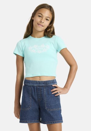 Mädchen mit langen Haaren, das ein hellblaues kurzes T-Shirt mit floralem Muster und dunkelblaue Jeansshorts trägt, posiert mit einer Hand auf der Hüfte.