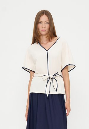 Frau trägt eine weiße V-Ausschnitt-Bluse mit marineblauem Besatz und Schleifengürtel, kombiniert mit einem marineblauen Rock, vor weißem, schlichtem Hintergrund stehend.