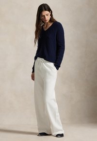 Femeie cu păr lung și închis la culoare, purtând un pulover bleumarin cu torsade, pantaloni albi cu croiala largă și pantofi negri, în picioare pe un fundal simplu bej.