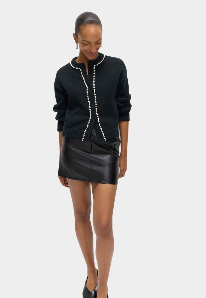 Cárdigan negro de punto con botones similares a perlas en el frente, combinado con una minifalda de cuero negro ajustada. Textura suave y un ajuste casual.
