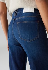 Dunkelblaue Denim-Jeans mit hoher Taille, ausgestattet mit orangen Nähten und zwei Gesäßtaschen. Der Bund zeigt ein weißes Markenetikett.