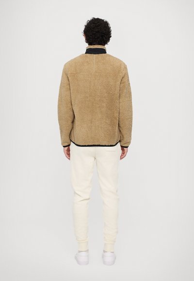 Tan fluffy fleece jacket with a black collar, seen from the back. White sweatpants and shoes complete the outfit. Minimalist design.