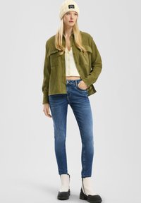 Chaqueta de camisa de pana oliva con dos bolsillos en el pecho, top corto de punto blanco, vaqueros ajustados azules y botas de tobillo con plataforma negras.