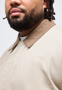 Κοντινό πλάνο ενός άνδρα με πλήρη γενειάδα που φοράει ένα μπεζ μπουφάν με υφασμένο καφέ γιακά από κορντούρο.