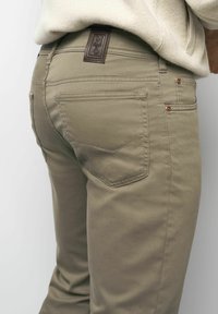 Close-up van een persoon die beige broek draagt met een achterzak en een leren merketiket met de tekst "M5" op taillehoogte.