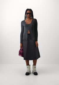 Black ribbed long-sleeve top with front slit, paired with a quilted black skirt. Silver and black knee-high boots, holding a shiny pink handbag.