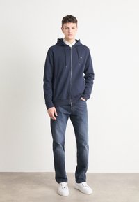Marine blauwe hoodie met rits, koordjes, frontzakken en een subtiele logo. Gecombineerd met donkere spijkerbroeken en witte sneakers. Vlotte textuur.