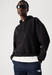 Zwarte cropped hoodie met trekkoord, zachte textuur, gedragen over een wit shirt, gecombineerd met marineblauwe broek met een logo. Zwarte zonnebril maakt de look compleet.