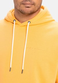 Yellow hoodie made of soft fabric, featuring a front pocket, white drawstrings with metal ends, and embroidered logo detail.