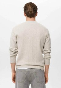 Hombre con un suéter de canalé beige, con las mangas arremangadas, y jeans grises, visto desde atrás sobre un fondo blanco liso.