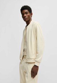 Lichtbeige bomberjack met een gladde textuur, voorzien van een ritssluiting en zijzakken, gedragen over een bijpassend T-shirt met ronde hals.