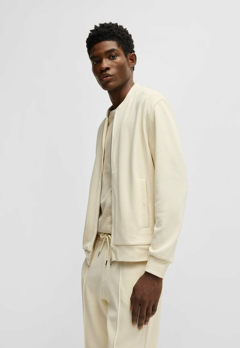 Lys beige bomberjakke med en glat tekstur, der har en lynlåslukning og sidelommer, båret over en matchende crew neck t-shirt.
