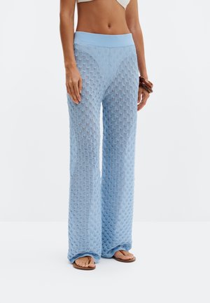 Kvinde iført lyseblå, teksturerede bukser med vide ben og høj talje, kombineret med brune sandaler og en hvid crop top.