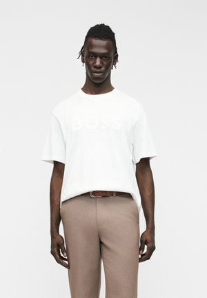 Άνδρας με πλεγμένα μαλλιά, φορώντας λευκό υπερμεγέθες T-shirt με διακριτικό κείμενο και μπεζ παντελόνι, στέκεται μπροστά σε απλό φόντο.