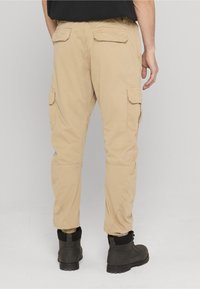 Bēšas kravas bikses ar sānu kabatām, elastīgām aprocēm un taisnu piegriezumu. Valkātas ar melniem zābakiem, no aizmugures redzamas šuvju detaļas.