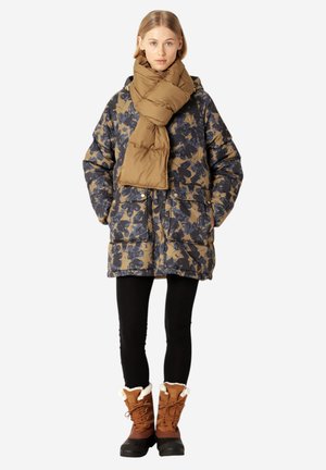 Pufferjack met marineblauw en tan bloemmotief, tan sjaal, zwarte legging en bruine laarzen met bontvoering tot de enkel. Vier buitenzakken op de jas.