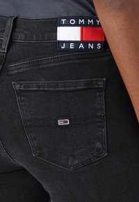 Černé džínové kalhoty s zadní kapsou, na které je malý vyšívaný motiv v červené, bílé a modré barvě. Materiál má hladkou texturu.