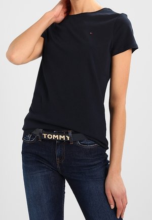Kvinde iført en ensfarvet sort kortærmet t-shirt og mørkeblå jeans med sort bælte, der viser "TOMMY" med store bogstaver.