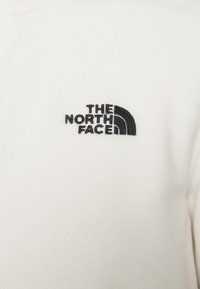 Valge fliisdress, millel on must broderitud logo "The North Face", kusjuures disain on kaarekujuline ja õlavarre piirkonnas on ümarad aktsendid.