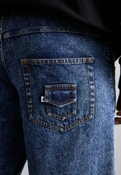Blue denim jeans with a large back pocket featuring orange stitching and a smaller pocket overlay. Fabric has a textured finish.