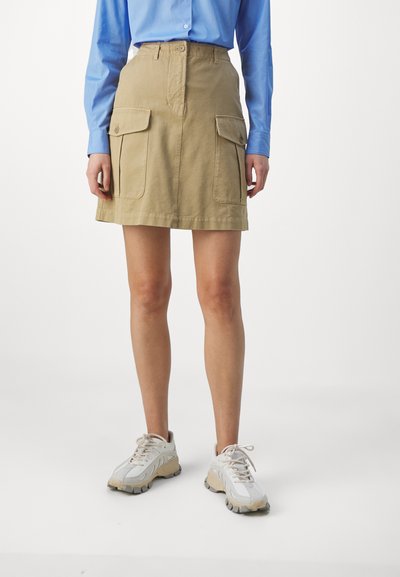 Gonna cargo khaki con due grandi tasche laterali, chiusura a bottone e silhouette dritta, abbinata a una camicia azzurro chiaro con bottoni e a sneaker bianche.