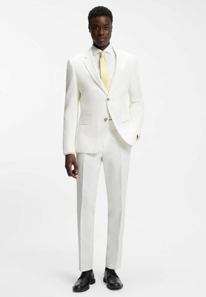 Mann trägt einen maßgeschneiderten weißen Anzug mit gelber Krawatte und schwarzen Anzugschuhen, steht vor einem schlichten weißen Hintergrund.