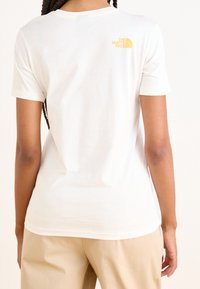 Camiseta blanca de algodón con un pequeño logo amarillo en la parte superior de la espalda, mangas cortas y un diseño clásico de cuello redondo.