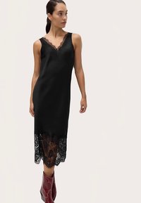 Zwarte mouwloze jurk met een kanten zoom en een v-hals, uitgevoerd in een gladde stof. Model draagt rode laarzen voor contrast.