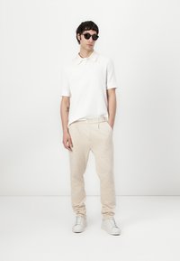 Hvit polo shirt, beige mønstrede bukser og hvite joggesko. Modell med svarte solbriller, står foran en ensfarget bakgrunn.