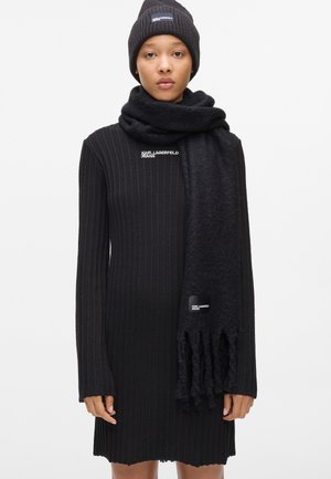 Vestito nero a coste con maniche lunghe, accompagnato da un berretto in maglia e una sciarpa coordinati. La sciarpa presenta bordi frangiati e un dettaglio con il logo.