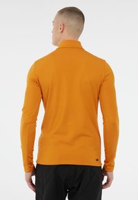 Vyras, atsigręžęs atgal, vilkintis prigludusią ilgomis rankovėmis garstyčių geltonumo marškinių ir juodas kelnes priešais paprastą baltą foną.