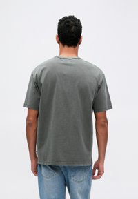 Šedé bavlnené tričko s krátkymi rukávmi, okrúhlym výstrihom a voľným strihom, v kombinácii s svetlomodrými džínsami. Zadný pohľad ukazuje minimálne detaily.