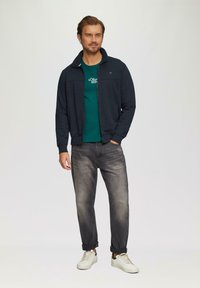 Marineblaues Zip-Jackett über einem grünen T-Shirt, kombiniert mit ausgebleichten schwarzen Jeans und weißen Turnschuhen. Merkmale sind ein Stehkragen und eine legere Passform.