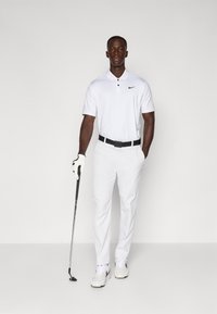 Vit kortärmad polo med en svart swoosh, vita byxor, svart bälte och vita sportskor. Håller en golfklubba.