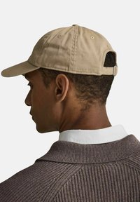 Beige bomullscap med en böjd skärm, justerbart band och logodetalj bak. Bärs med en brun ribbad tröja och vit krage.