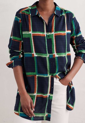 Koszula na długi rękaw zapinana na guziki w kratkę w kolorach marynarskiego granatu, zieleni, bieli, pomarańczu i błękitu, noszona z jasnymi spodniami.