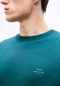 Άνδρας που φοράει πετρόλ φούτερ με στρογγυλή λαιμόκοψη και το λογότυπο "Mads Nørgaard Copenhagen" στο στήθος, κοιτάζοντας αριστερά, φωτεινό φόντο.