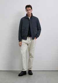 Man wearing a dark blue jacket over a black sweater, beige pants, black belt, and black shoes standing against a plain light wall.