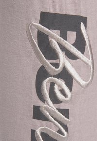 Geborduurd textiel met een zachte roze achtergrond. Kenmerkt grote zwarte letters en witte textuurelementen, met unieke stikdetails.