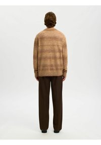 Personne aux cheveux mi-longs portant un pull marron dégradé, un pantalon large marron foncé et des chaussures noires, debout de dos.