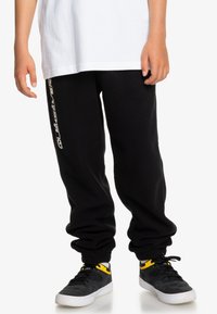 Svart träningsbyxor i mjukt tyg med elastiska muddar, med en vit logotryck på vänster sida. Kombineras med svarta sneakers.
