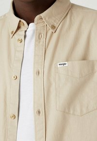 Camicia beige con bottoni, realizzata in cotone, caratterizzata da una tasca sul petto con etichetta, bottoni dorati e una vestibilità comoda.