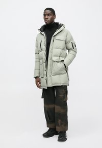 Ανοιχτό γκρι μπουφάν με κουκούλα, πολλαπλές τσέπες και μαύρες λεπτομέρειες, σε συνδυασμό με σκούρο καφέ παντελόνι cargo και μαύρα αθλητικά παπούτσια.