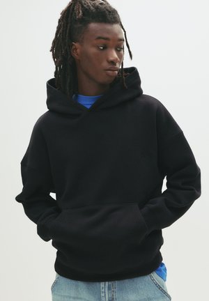 Schwarzer Kapuzenpullover mit geräumiger Kapuze, Fronttasche und gerippten Bündchen. Über einem blauen Hemd getragen, das ein dezentes Logo-Detail aufweist. Lockere Passform.