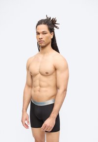 Άνδρας χωρίς πουκάμισο με dreadlocks στέκεται κοιτώντας μπροστά φορώντας μαύρα μποξεράκια Calvin Klein σε απλό ανοιχτό φόντο.