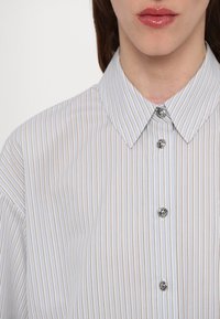 Gros plan d’une personne portant une chemise de couleur claire avec de fines rayures verticales et des boutons décoratifs brillants, col visible, lèvres partiellement montrées.