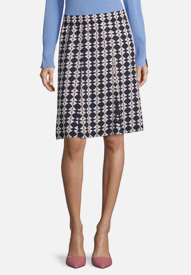 Laivastonsininen hame, jossa on valkoisia ja vaaleanpunaisia geometrisia kuvioita, pliseerattu design ja joustava vyötärönauha, yhdistettynä vaaleanpunaisiin teräviin korkokenkiin.