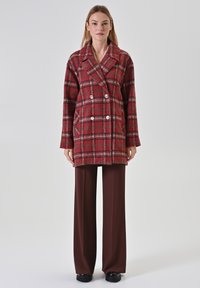Donna con un cappotto doppiopetto a quadri rossi, pantaloni larghi marroni e scarpe nere, in piedi davanti a uno sfondo chiaro e semplice.