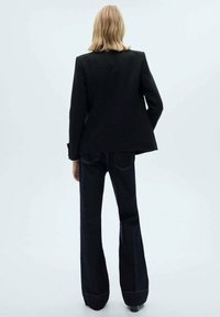 Sacou negru croit, cu umeri structurați, asortat cu pantaloni evazați din denim închis, cu cusături contrastante și o textură netedă.