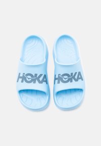Ζευγάρι ανοιχτό μπλε γλιστρητών από καουτσούκ της μάρκας Hoka με υφή στο πέλμα και σκούρο μπλε λογότυπο στο λουράκι, φαίνεται από ψηλά.
